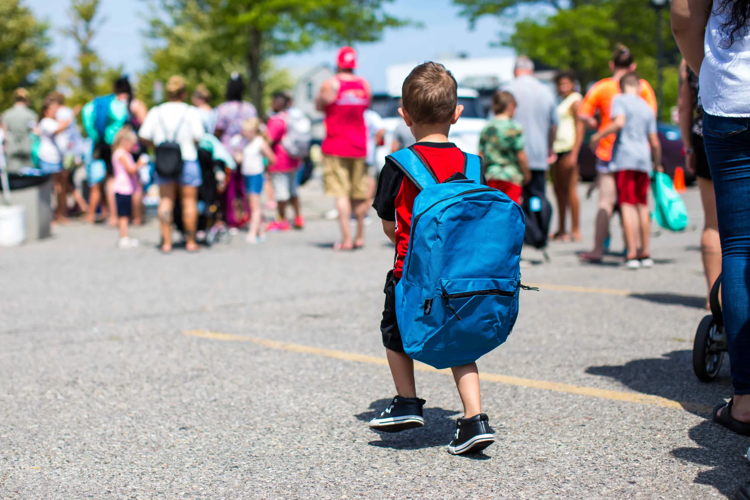 Young boy walks with a blue backpack towards a small crowd of children