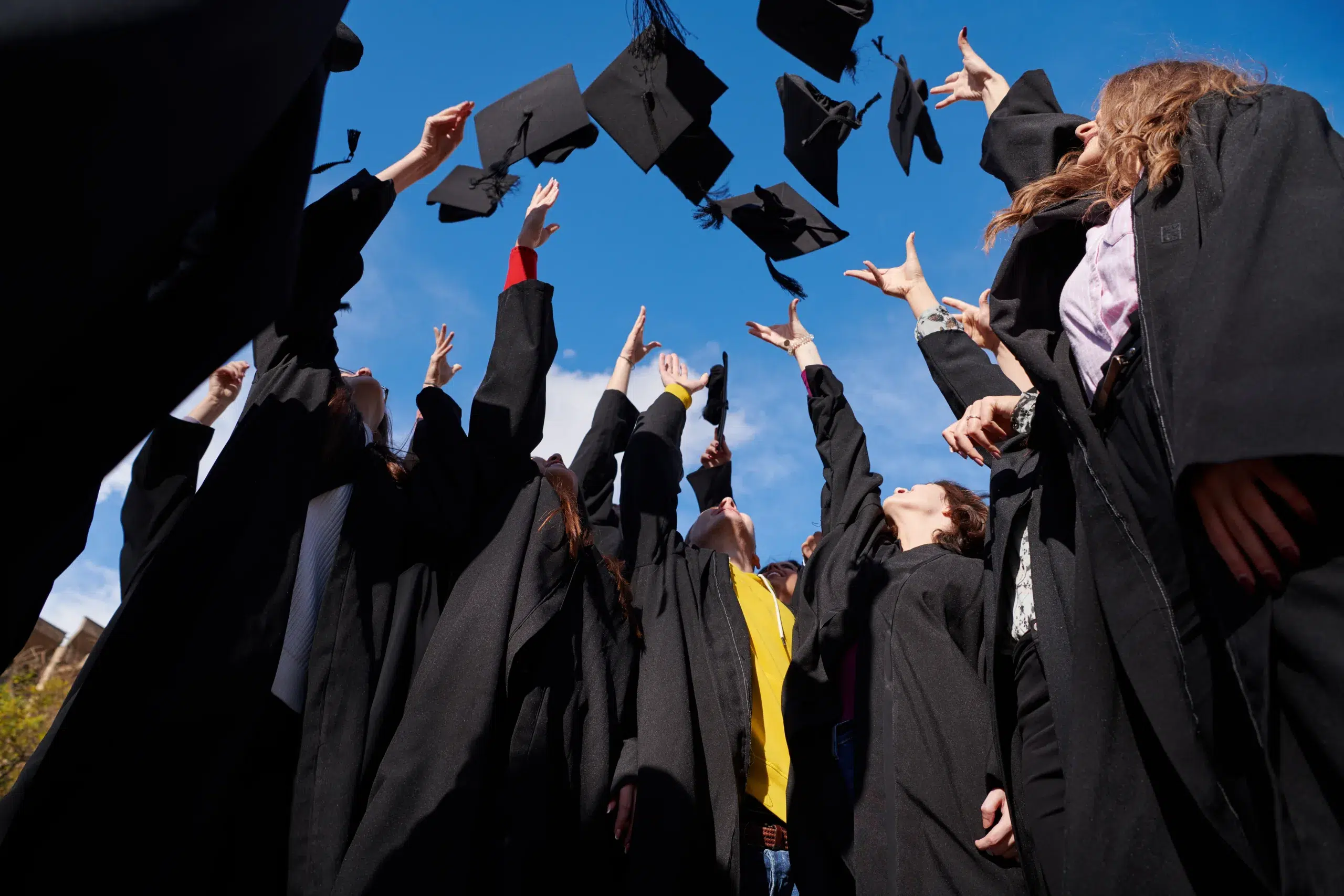 Congratulations Low angle view of happy group of six young cheerful graduates in black gowns, throwing up their head wear in the air and celebrating, in blue summer sky, laughing, enjoying - Community Choice Credit Union Michigan
