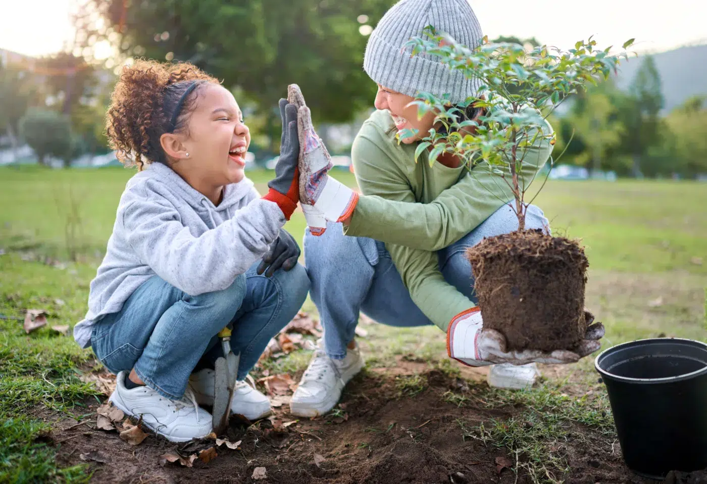 High five, child and woman with plant for gardening at park with trees in nature garden environment. Happy volunteer family planting for growth, ecology and sustainability for community. Community Choice Credit Union in Michigan